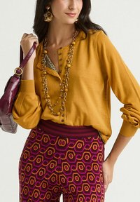 Pull jaune moutarde avec col boutonné, associé à un pantalon bordeaux à motifs hexagonaux roses et orange.