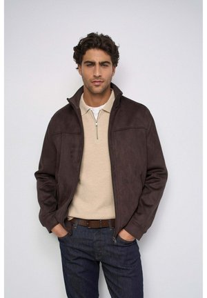 Uomo che indossa una giacca marrone scuro sopra una camicia beige con zip e jeans scuri con le mani in tasca su sfondo semplice.