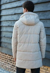 Graue Steppjacke mit Kapuze, die gesteppten Abschnitte und eine weiche Textur aufweist. Entworfen für Wärme, von hinten an einer Holzwand präsentiert.