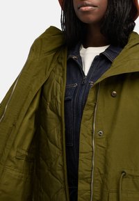 Olivgrön quiltad jacka med dragkedja och knapplås, i kombination med en mörk denimoverall. Texturerat tyg och funktionella designfunktioner.