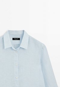 Hellblaues Leinenhemd mit langen Ärmeln, spitzem Kragen und drei weißen Knöpfen vorne, flach auf einem weißen Hintergrund aufgelegt.