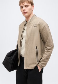 Νέος άνδρας που φορά μπεζ μπουφάν BOSS, ανοιχτό πουκάμισο, μαύρο παντελόνι, κρατώντας μια μαύρη τσάντα duffel με το ένα χέρι στην τσέπη.