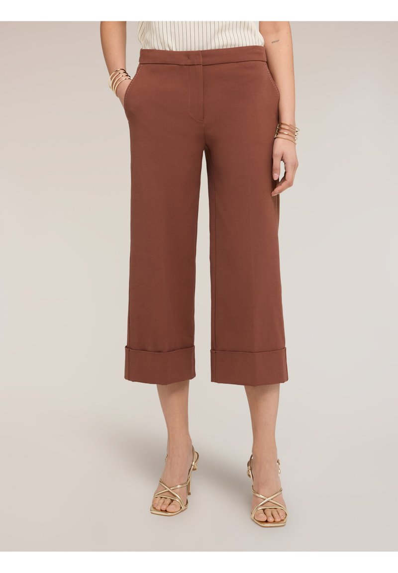 Kvinde iført brune, korte, vidde bukser og gyldne, snørede hælsandaler, stående med hænderne i lommerne på beige baggrund.