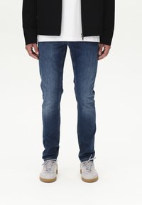 Modri denim kavbojke s prilegajočo zasnovo, rahlo zbledelimi stegni in ravnim krojem, v kombinaciji s svetlo sivimi supergami.