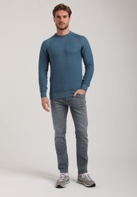 Camicia a maniche lunghe blu con texture e polsini a costine, abbinata a jeans grigi chiari e scarpe da ginnastica grigie. Il modello è in piedi su uno sfondo neutro.