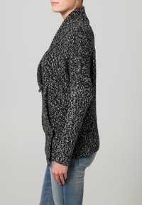 Cardigan tricoté en noir et blanc avec un motif texturé, un col châle et des accents de boutons. Coupe ample et manches longues.