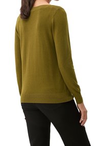 Kobieta ubrana w oliwkową długą swetrową bluzkę i czarne spodnie, pokazana z tyłu na tle białym.