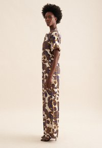 Abito lungo, aderente in crema con motivi floreali blu e oro che si avvolgono. Maniche corte a palloncino e vita cinte. Indossato con tacchi neri.