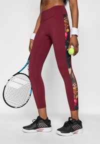 Burgundi, magas derékú leggings fekete virágmintás oldalsó panelekkel, nedvességelnyelő anyagból, fekete sportcipővel és teniszlabdával párosítva.