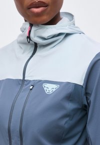 Κοντινό πλάνο ενός ατόμου που φοράει μπλε και ανοιχτό γκρι μπουφάν με ψηλό γιακά, φερμουάρ μπροστά και λογότυπο με ζωάκι στο στήθος.