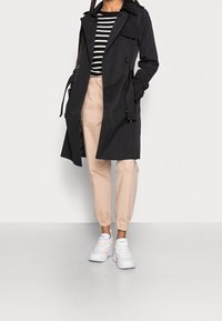 Schwarzer Trenchcoat mit doppelreihigen Knöpfen, beigen Jogginghosen und weißen Sneakers. Gestreiftes Oberteil in Lagenoptik mit Rundhalsausschnitt.