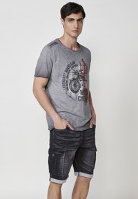 Camiseta gráfica gris con estampado de motocicleta y pantalones cortos negros de cargo con puños enrollados y bolsillos laterales. Tela de algodón suave.