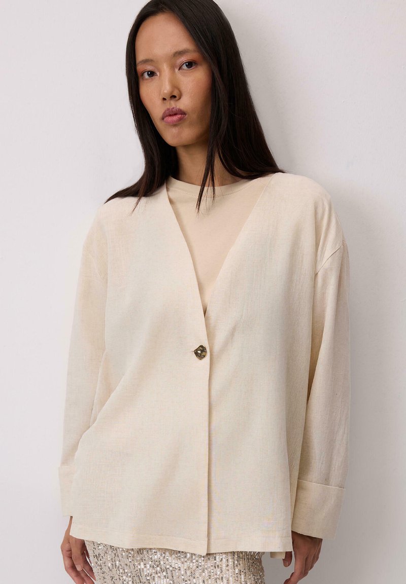 Sacou din in bej, cu decolteu în V, croială supradimensionată și un singur nasture decorativ. Dispune de mâneci lungi și o textură netedă.