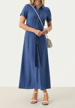 Kobieta stojąca, ubrana w niebieską sukienkę maxi z krótkim rękawem, zapinaną na guziki, z paskiem w talii, białe sandały na kostce oraz nosząca małą plecioną torebkę na ramię.
