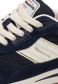 Gros plan d'une basket bleu marine et blanche avec des lacets couleur crème et le logo "Tamaris" sur le côté et sur l'étiquette de la languette.