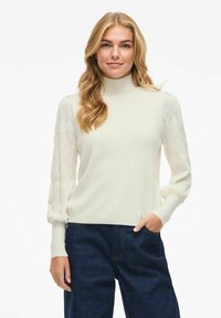 Mujer rubia vestida con un suéter blanco de cuello alto con mangas abullonadas y un patrón, y unos jeans de tiro alto azul oscuro, con una mano en el bolsillo.