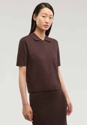 Femme aux cheveux lisses et foncés portant un haut zippé à manches courtes de couleur brun foncé et une jupe assortie, sur un fond clair uni.