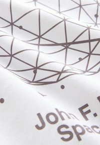Valkoinen kangas, jossa on geometrinen kuvio, jossa on risteäviä ruskeita viivoja ja pieniä ympyröitä. Teksti sisältää "John F. Kennedy" ja "Avaruus."