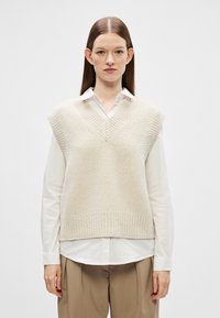 Colete tricotado bege com decote em V e design sem mangas, sobre uma camisa branca com colarinho. Textura suave com detalhes em barra canelada.