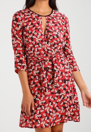 Woman wearing a red floral dress with white flowers, three-quarter sleeves, a front keyhole neckline, and a waist tie belt.