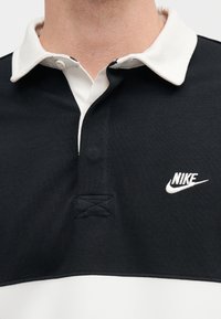 Μαύρο και λευκό πουκάμισο πόλο Nike με λευκή γιακά, κουμπωτό ντεκολτέ και κεντημένο λογότυπο Nike στο στήθος.