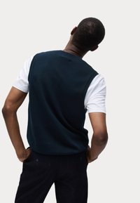 Gilet a maglia blu navy con collo rotondo sopra una maglietta bianca, abbinato a pantaloni scuri. Presenta una texture liscia e orlo a costine.