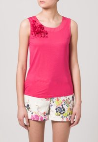 Roze mouwloze top van soepel materiaal, met een geplooide halslijn en een bloemaccent op de linkerschouder. Gepaard met bloemen shorts.