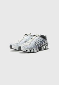 Nike Shox sneakers in metallic silver and black, featuring a structured upper design, flexible sole with visible coil springs, and laces.