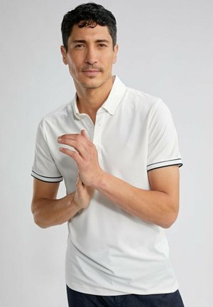 Homme aux cheveux courts et foncés, portant un polo blanc avec une bordure noire, les mains jointes devant un fond clair uni.