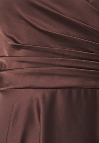 Καφέ σατέν ύφασμα με απαλές πτυχώσεις, που διαθέτει κατακόρυφες πτυχές στη μέση, δημιουργώντας μια λεία υφή και κομψό σιλουέτα.