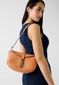 Sac à main en cuir texturé couleur cognac avec ferrures dorées et fermeture magnétique. Présente une forme courbée et une sangle amovible pour un port polyvalent.
