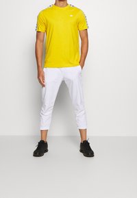 Camiseta amarilla de manga corta con logo, combinada con pantalones cortos blancos. Zapatillas deportivas negras completan el atuendo. Diseño simple y casual.