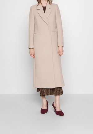 Manteau long en laine beige clair avec un col à revers, poches avant et silhouette épurée, porté sur une robe à carreaux marron et des talons bordeaux.