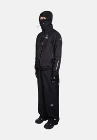 Tenue d'extérieur noire comprenant une veste à capuche et un pantalon ample. Le matériau semble durable. Comprend des gants et des détails de marque distinctifs.