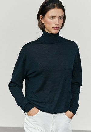 Donna che indossa un maglione dolcevita nero e pantaloni bianchi con le mani in tasca, in piedi contro uno sfondo chiaro e uniforme.