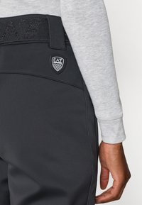 Μαύροι φόρμες με υφασμάτινη ζώνη μέσης με υφή που φέρει το λογότυπο "EA7". Λείο ύφασμα και λεπτομερής ραφή ορατή στο πλάι και πίσω.