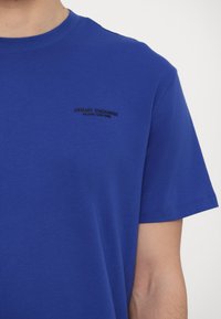 Μπλε βαμβακερή μπλούζα με κοντά μανίκια και στρογγυλή λαιμόκοψη. Ένα μαύρο λογότυπο είναι τυπωμένο στην αριστερή πλευρά του στήθους.