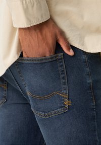 Jean en denim bleu foncé avec des coutures jaunes. Une main est placée dans la poche arrière. Les jeans présentent un design de poche incurvée.