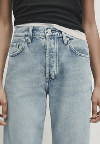 Niebieskie dżinsy z wysokim stanem, z przodu z guzikami i kieszeniami, noszone z warstwowymi czarnymi i białymi topami, ręce swobodnie opadające wzdłuż ciała.