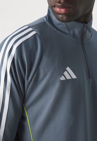 Gray athletic zip-up top with white stripes along the shoulders and a green accent. Features an embroidered logo on the chest.