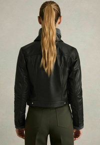 Schwarze Lederjacke mit Schulternähten, umgeschlagenem Kragen und silbernen Nieten, getragen über einem grauen Pullover und kombiniert mit olivgrünen Hosen.