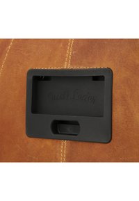 Brown leather surface with visible stitching. Black plastic slot with "Gusti Leder" embossed. Simple geometric design, smooth texture.