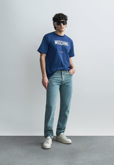 Tamnoplava pamučna majica s ispisima "MOSCHINO" i "Milano", uparena sa svijetloplavim trapericama i bijelim tenisicama. Model nosi velike tamne sunčane naočale.