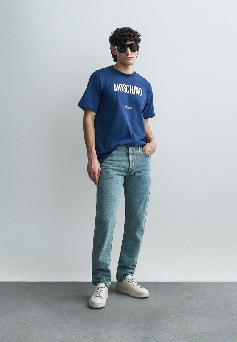 Marineblauw katoenen t-shirt met de teksten "MOSCHINO" en "Milano", gecombineerd met lichtblauwe jeans en witte sneakers. Het model draagt oversized donkere zonnebrillen.