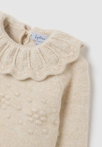 Strickpullover in Creme mit einem strukturierten Bubbelmuster, gewelltem Kragen und subtilen Glitzereffekten; Etikett am Hals sichtbar.