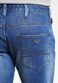 Blue denim jeans featuring a back pocket with a metal logo accent, contrasting stitching, and a classic fit. Textured and slightly faded.