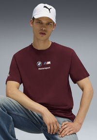 Mørkerød bomulds t-shirt med BMW Motorsport-logo og sorte detaljer, parret med lyseblå denimjeans og en hvid caps med et sort logo.