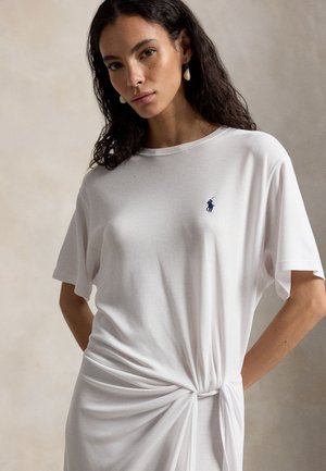 Kobieta z długimi ciemnymi włosami w białej, krótkiej sukience z węzłem po boku i małym haftowanym logo polo na piersi, stojąca na neutralnym tle.