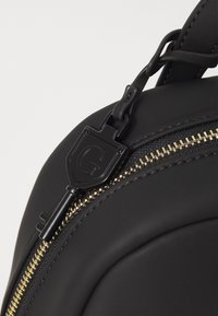 Sac à dos noir en matière texturée, présentant une fermeture éclair dorée bien visible et un tirette de fermeture zippée en relief avec le logo. Surface lisse avec des courbes subtiles.