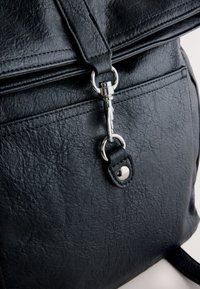 Gros plan d’un rabat de sac en cuir noir texturé sécurisé par une fermeture métallique argentée et un anneau avec un détail de sangle en cuir cousu.